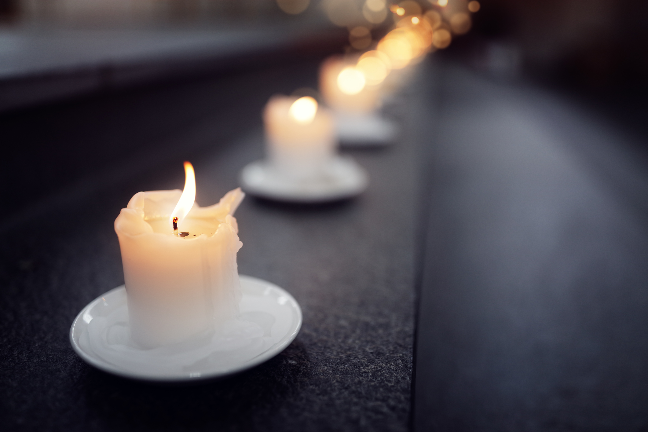Photo of several short, burning white candles in a line on a black surface blurring as the line continues into the background with the only the first candle and flame in sharp focus. 