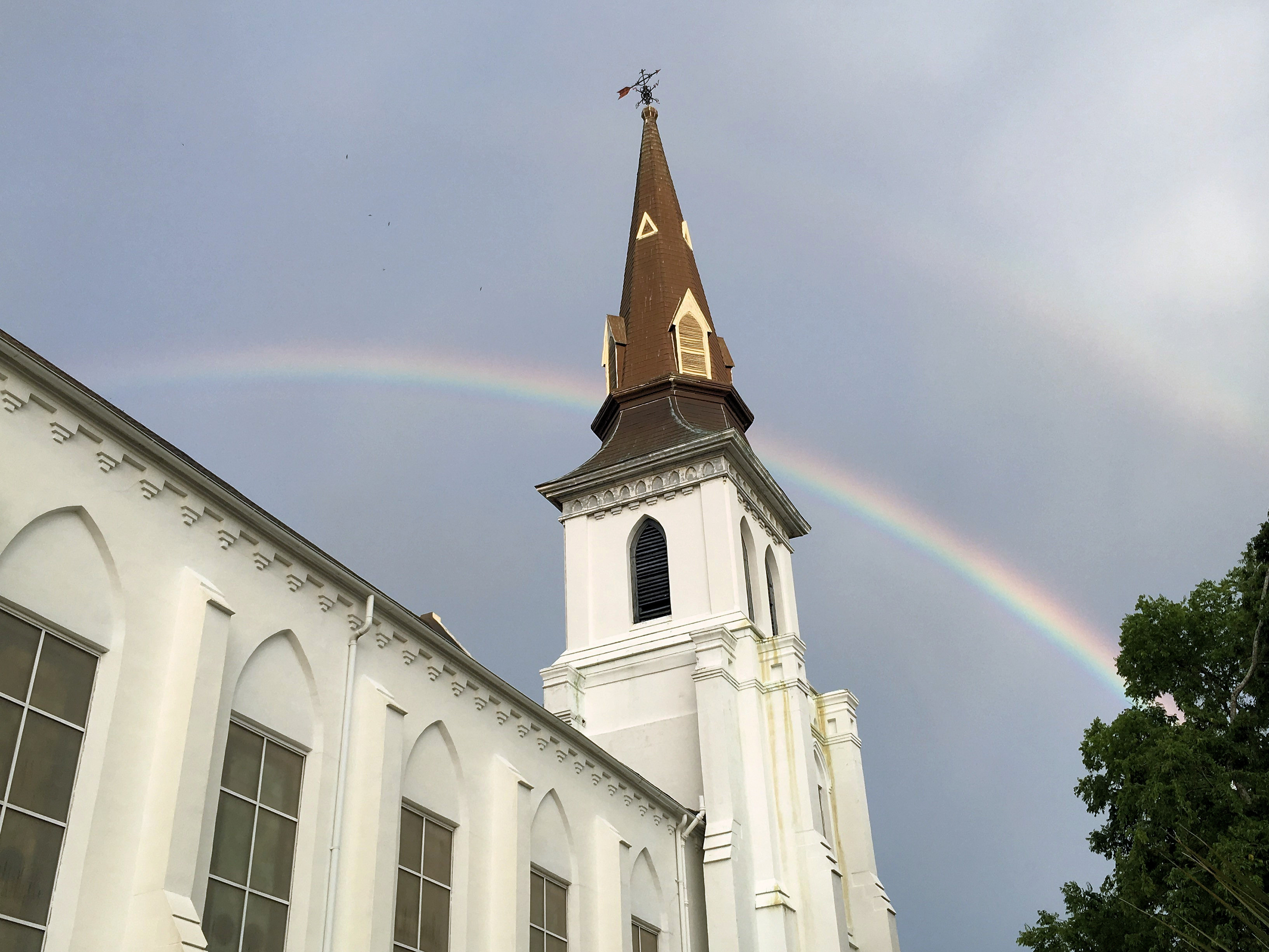 Photograph of the spire of the Mother Emanuel Church spire with a rainbow in the dark sky behind, taken at the memorial for victims of the mass shooting which occurred there. 