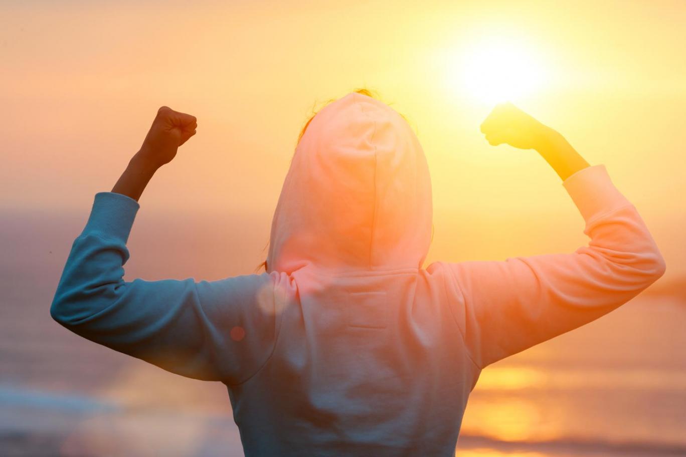 Image of the back of a person wearing a hoodie at sunset on a beach, their arms raised as if celebrating.